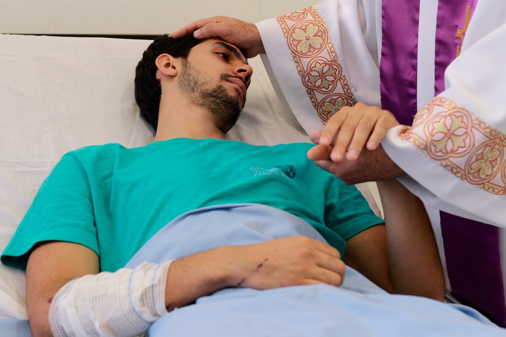 Sacerdote de estola roxa unge a testa de um homem doente deitado na cama